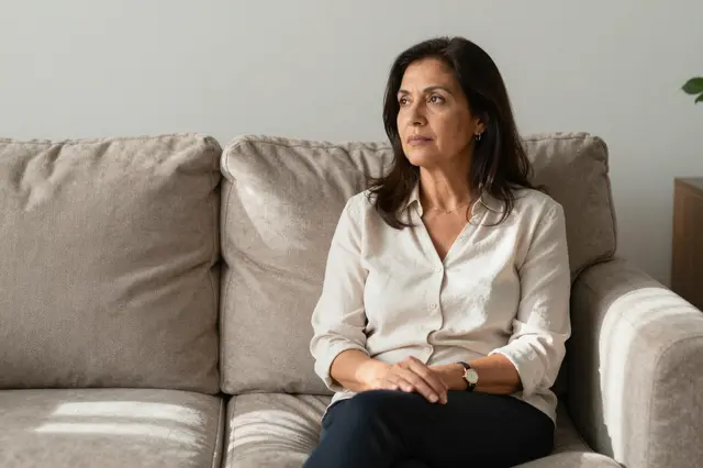 A thoughtful Latina woman in her sixties sits alone on a beige sofa in soft afternoon light.