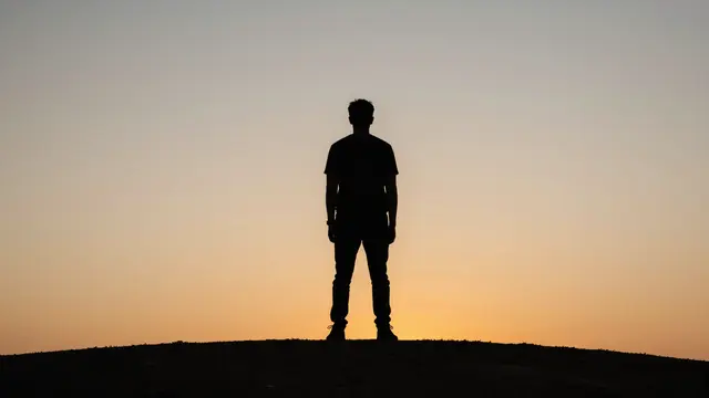 A dark silhouette of a standing person on a hill against a warm gradient sunset sky