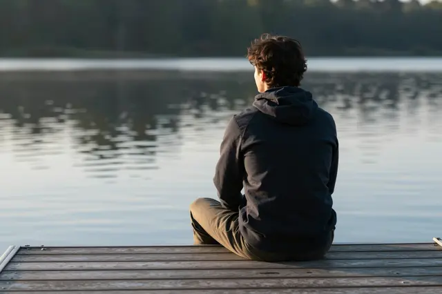 View from behind of a person sitting on a wooden dock overlooking a calm lake at dawn