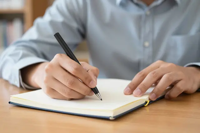 Close-up of an adult person's hands writing with a pen in an open lined notebook on a wooden table