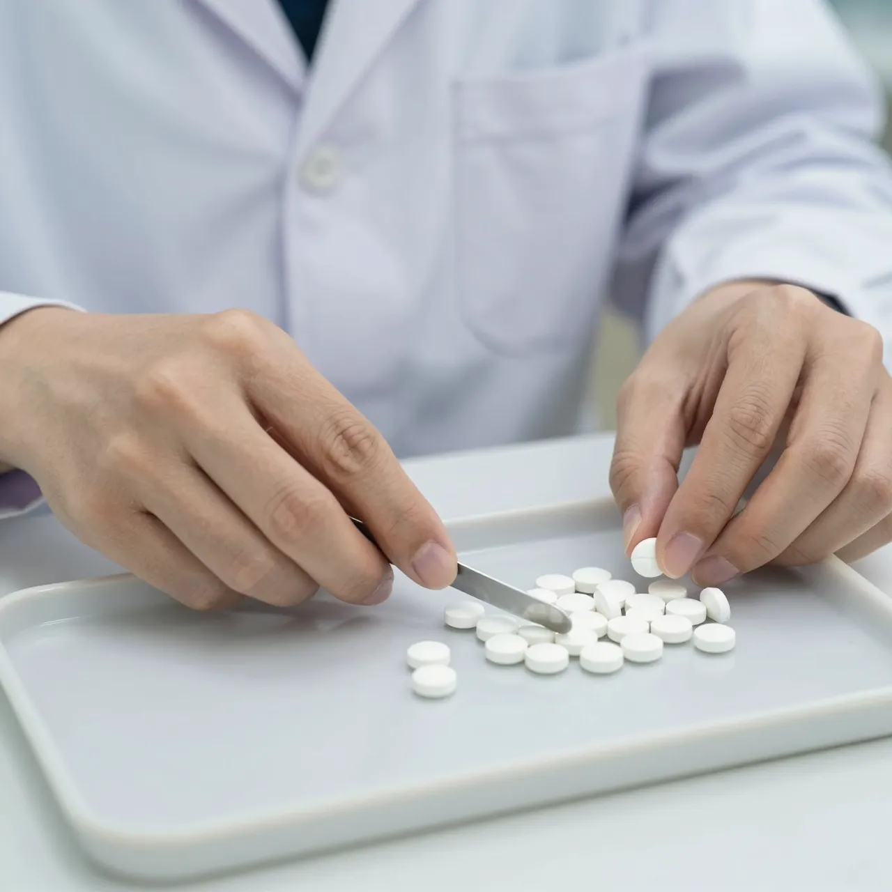 A pharmacist's hands count white tablets on a tray using a metal spatula