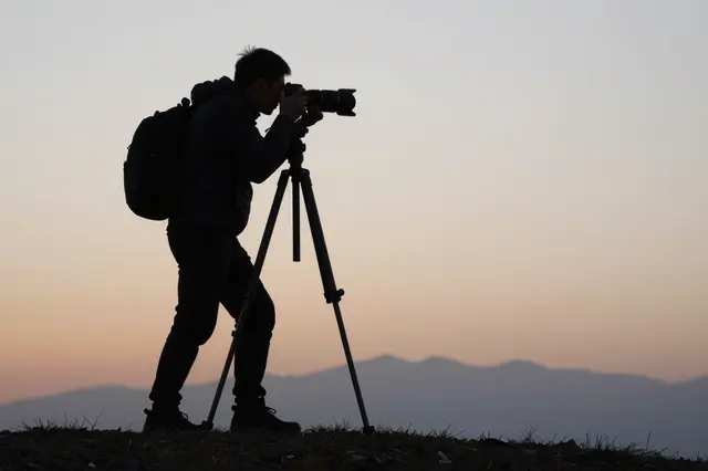 Silhouette of a photographer with a backpack using a camera on a tripod atop a grassy ridge at sunset