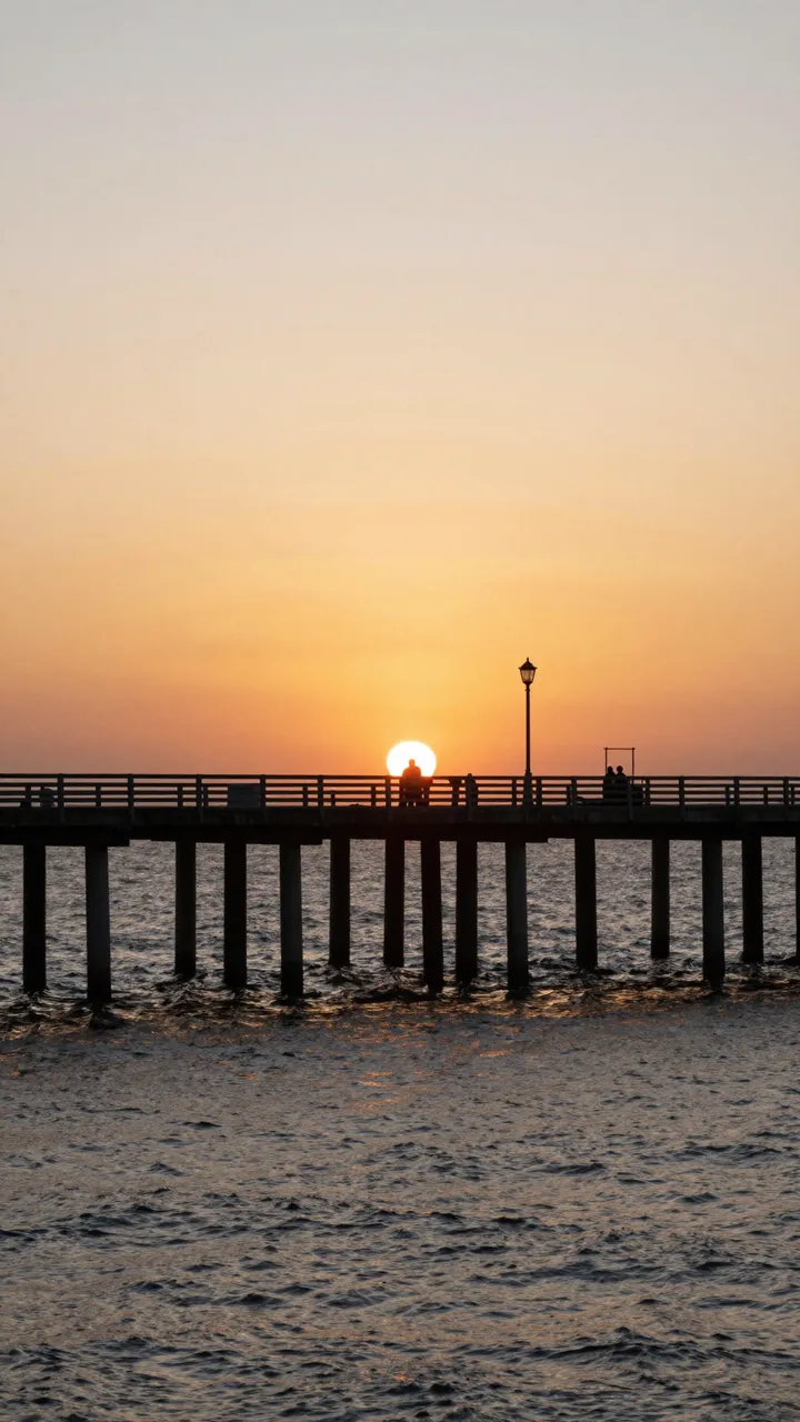 A silhouetted ocean pier stretches across choppy water beneath a warm orange and peach sunset sky with small visitor