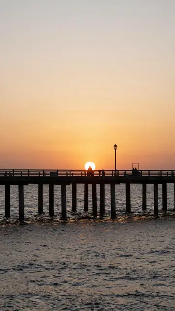 A silhouetted ocean pier stretches across choppy water beneath a warm orange and peach sunset sky with small visitor