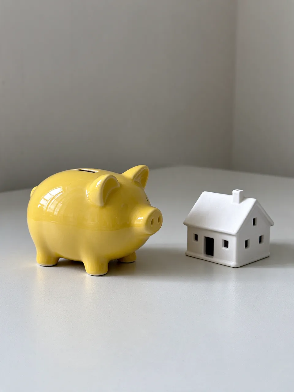 A glossy yellow ceramic piggy bank sits next to a small white house model on a pale gray table in soft daylight