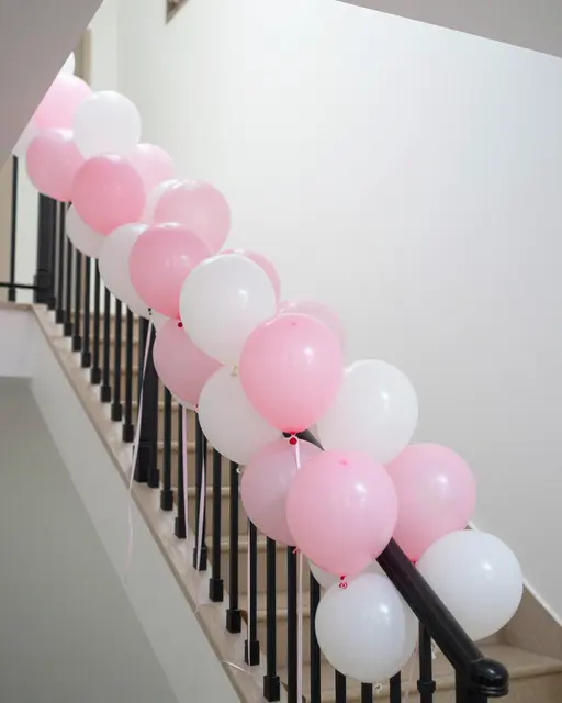 Pink and white balloons tied to a black stair banister with trailing ribbons in a bright neutral hallway.