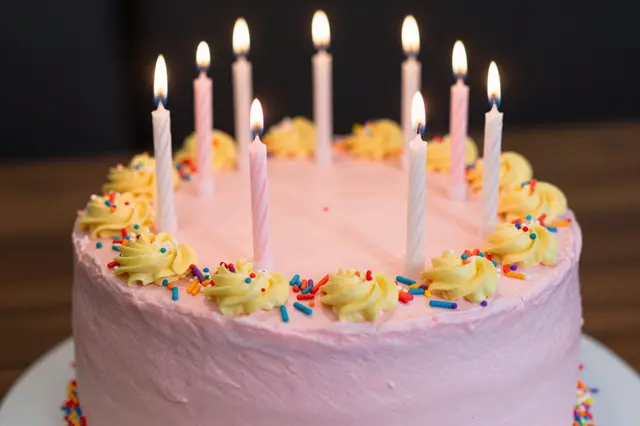 A pink frosted birthday cake topped with lit candles, yellow frosting swirls and colorful sprinkles.