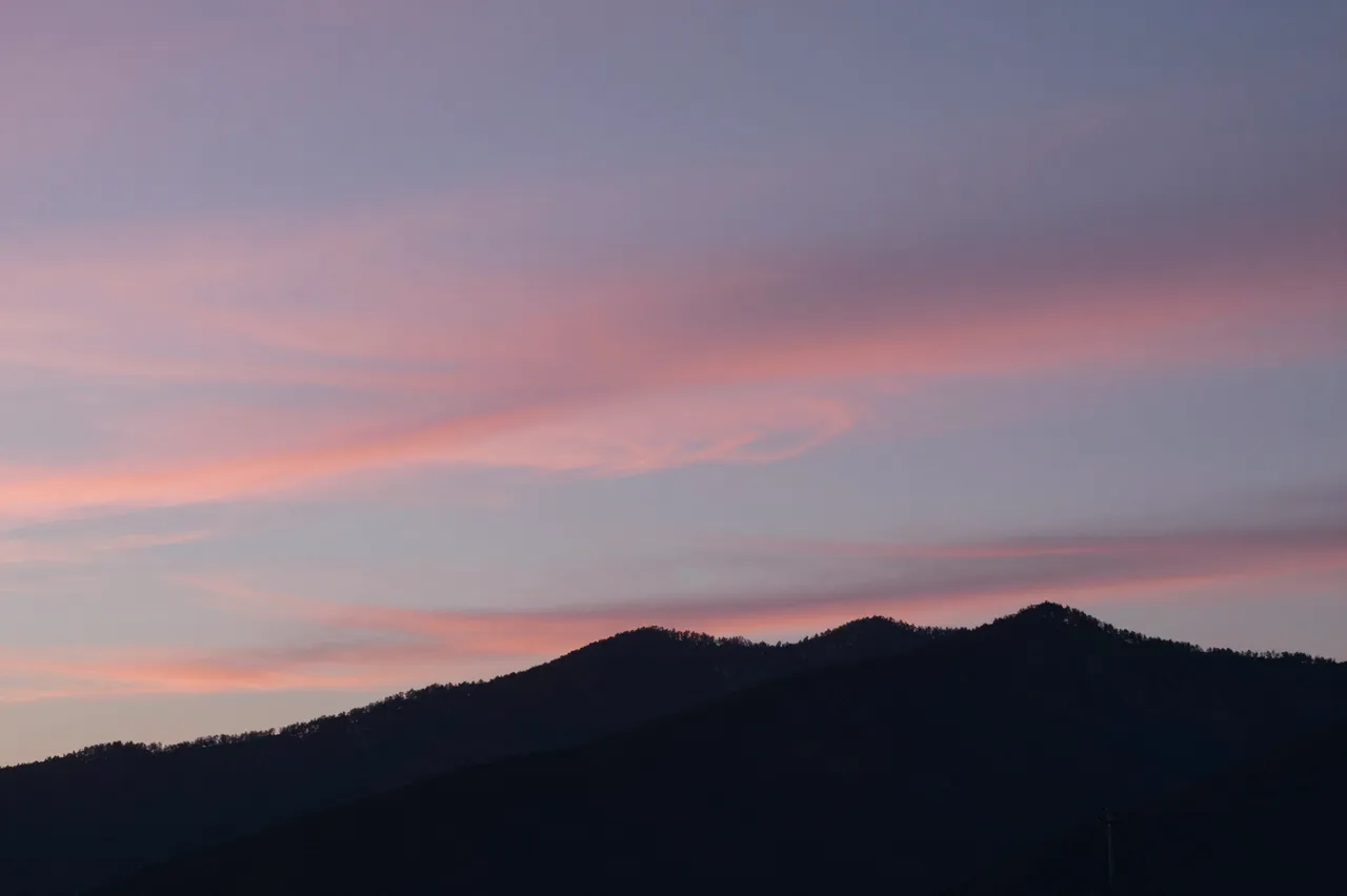 Soft streaked pink cirrus clouds drift above a dark mountain silhouette at early dawn