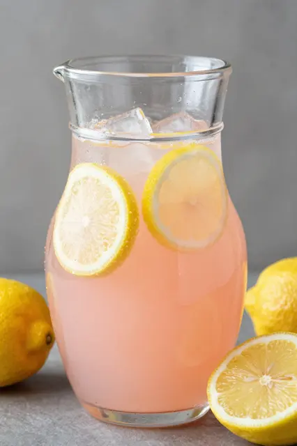 Glass pitcher of pink lemonade with ice and lemon slices, surrounded by fresh lemons on a gray surface