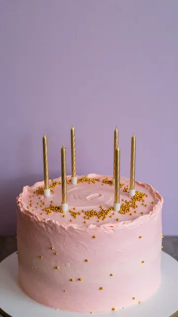 Pink ombre frosted birthday cake with gold sprinkles and thin candles against a lilac wall