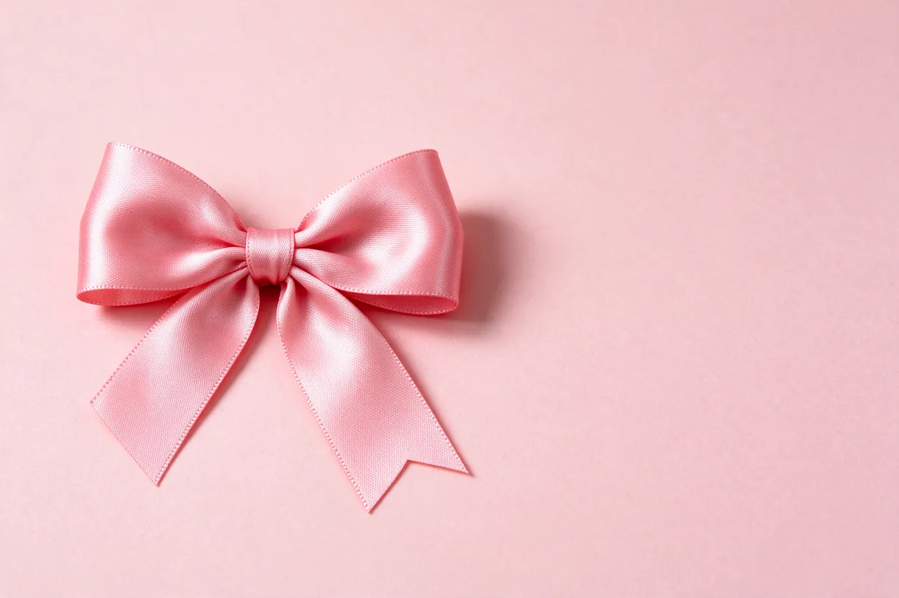 A neat pink satin ribbon bow sits on the left of a solid light pink paper backdrop with empty space to the right.