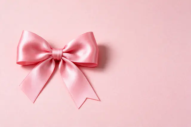 A neat pink satin ribbon bow sits on the left of a solid light pink paper backdrop with empty space to the right.