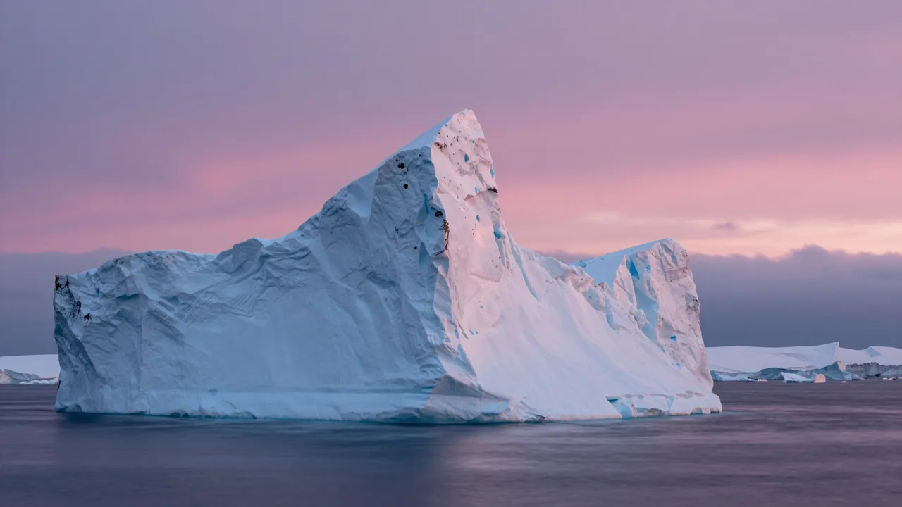 A large textured iceberg floats in calm dark polar waters beneath a soft pink and purple midnight sky.