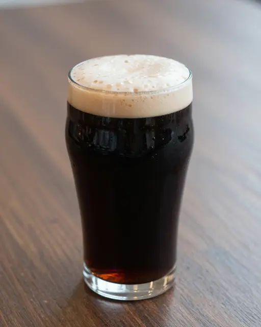 A glass of dark stout with a thick creamy foamy head sits on a wooden tabletop.