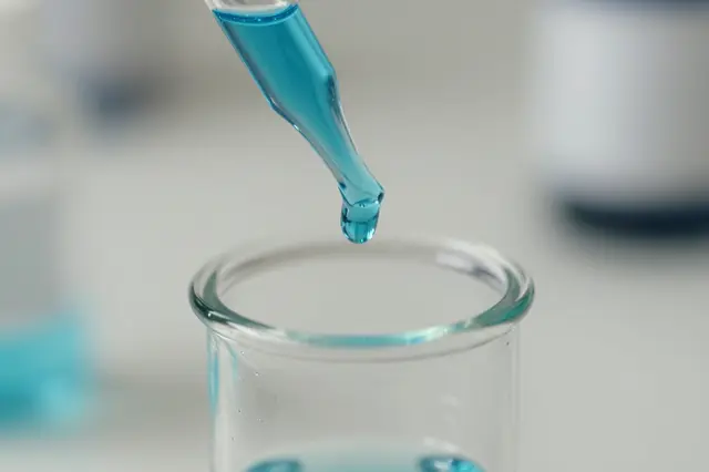 A macro close-up of a glass pipette releasing a drop of blue liquid above a clear lab vial.