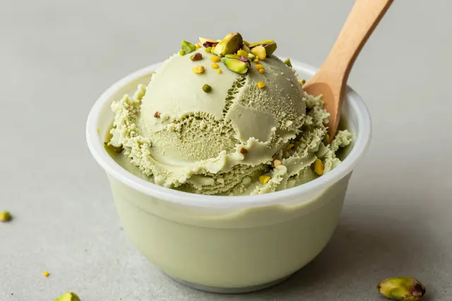 Scoop of pistachio gelato topped with nuts sits in a small cup with a wooden spoon on a neutral surface
