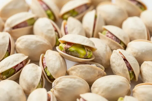 Dense close-up of shelled pistachios, many partially opened to show green nut kernels, filling the entire frame