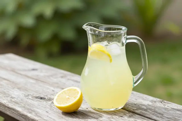 A glass pitcher of iced lemonade sits next to a halved lemon on a weathered wooden picnic table in a sunlit garden.