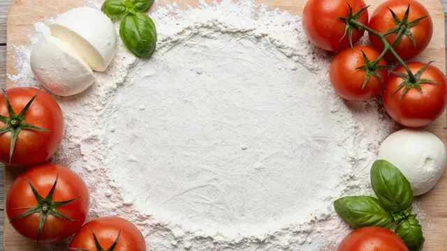 Top view of a floured wooden board bordered with tomatoes, mozzarella and fresh basil with central empty copy space.