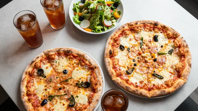 Overhead view of two pizzas, a fresh salad, and iced tea glasses arranged on a light table