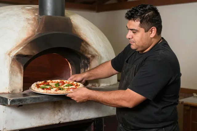 Heavyset pizzeria owner slides an uncooked pizza toward a lit wood-fired oven
