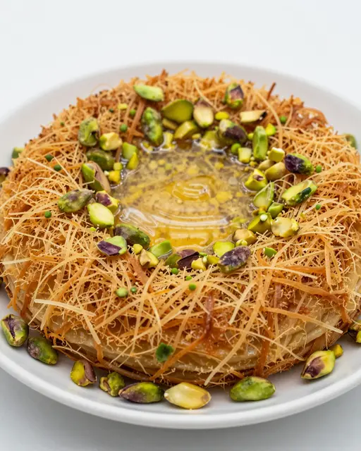 A close-up of golden crispy kunafa topped with chopped pistachios and glistening syrup on a white plate.