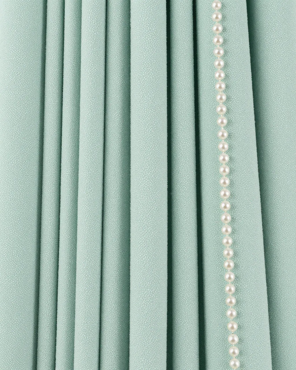 Full frame close-up of pleated pale mint shagreen surface paired with a straight strand of small glossy pearls
