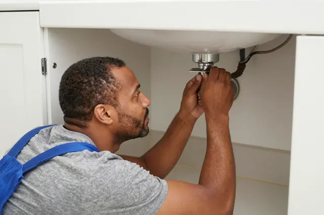 A male plumber in his forties works under a bathroom vanity to install or adjust a sink drain.