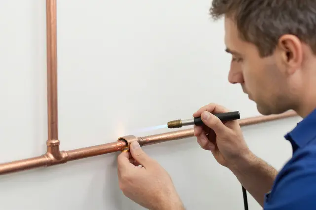 Side view of a male plumber using a small torch to solder a copper pipe joint on a plain wall