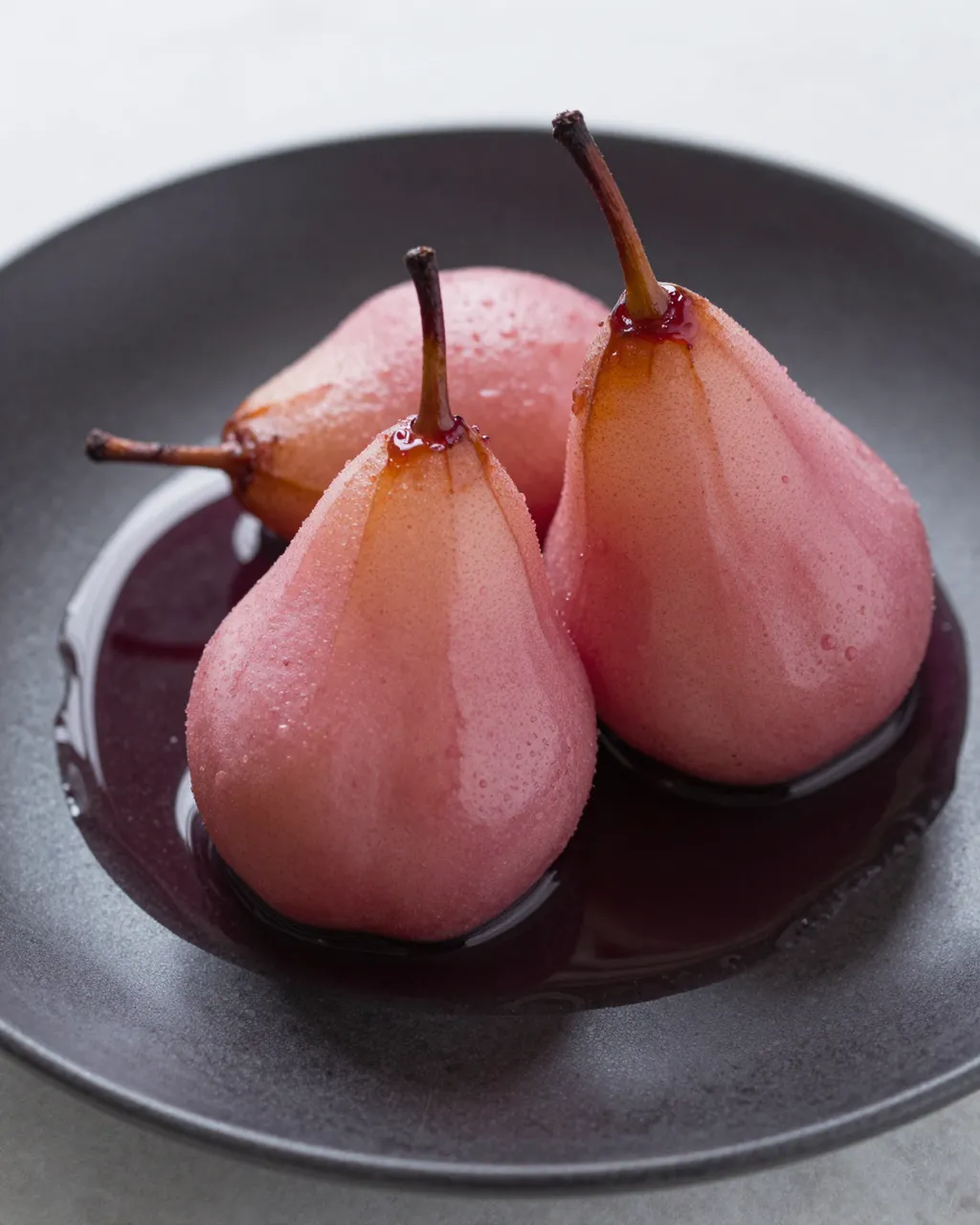 Three pink poached pears rest in red wine syrup on a dark matte plate