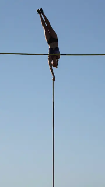 A female pole vaulter balances vertically at the top of her vault above the crossbar against a clear blue sky.