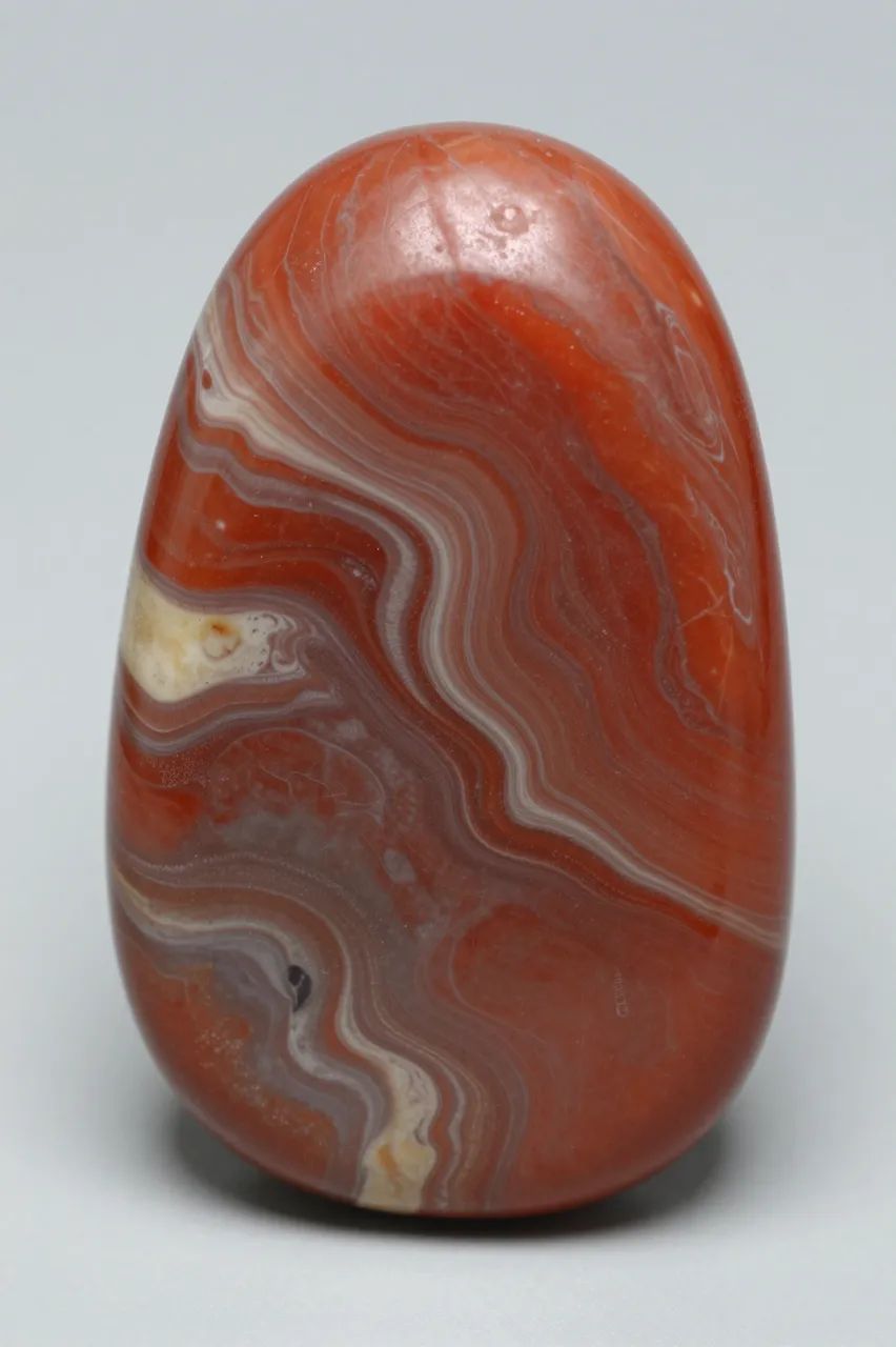 A smooth polished red jasper tumbled stone with swirling creamy and gray banding rests on a light neutral background