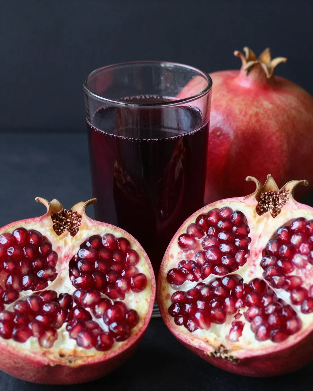 A glass of deep red pomegranate juice sits beside whole and halved pomegranates on a dark background.