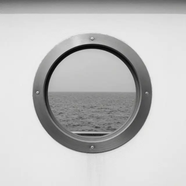 Black and white view through a circular metal porthole of calm open sea beyond a white wall