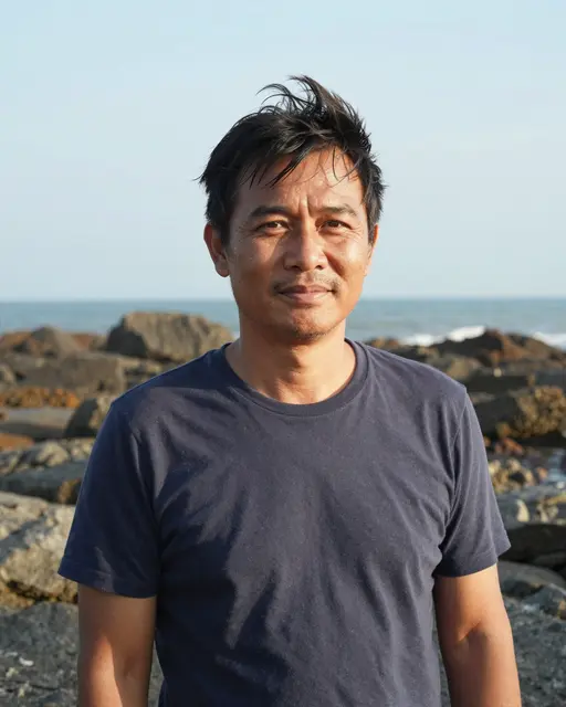 Middle aged Southeast Asian man with wind tousled hair stands on a rocky ocean coastline in bright afternoon sunlight