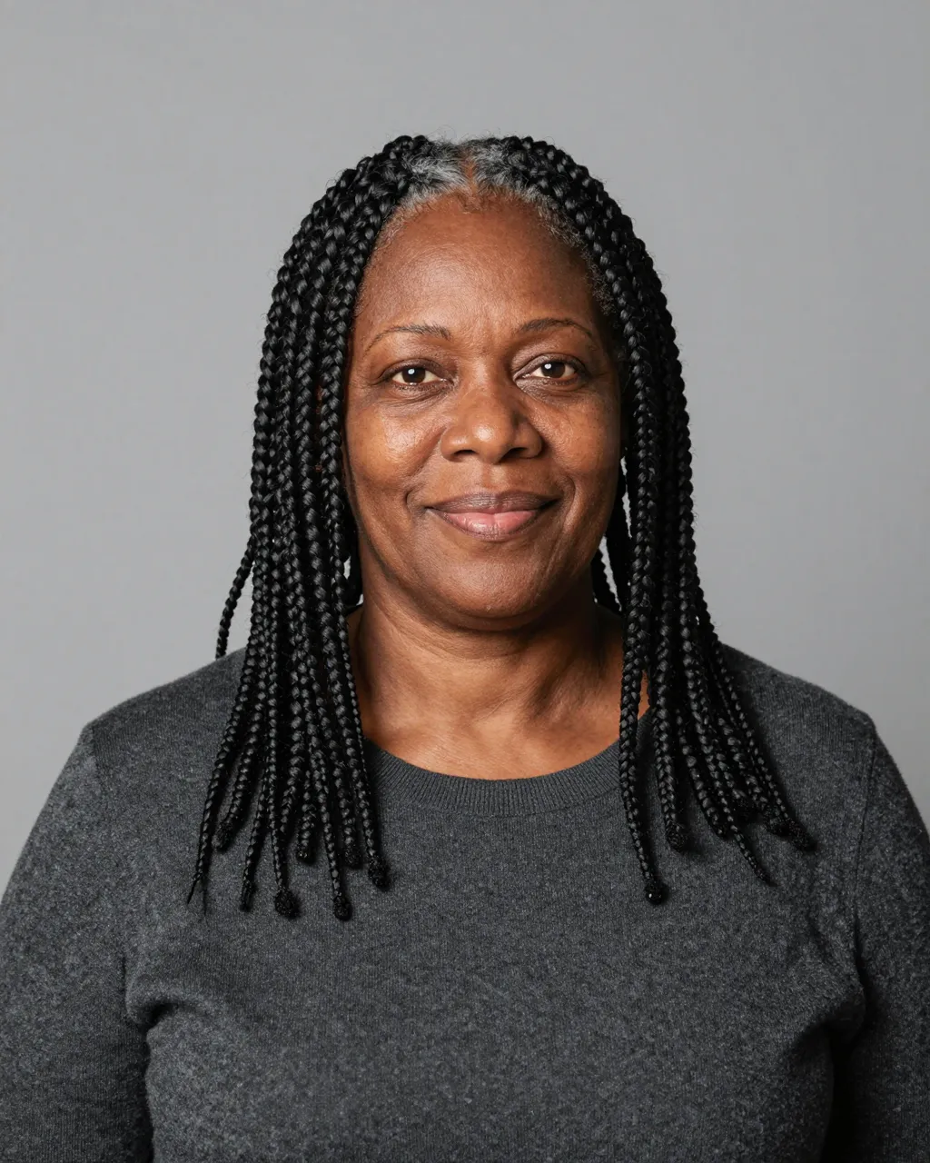 Studio portrait of a Black woman in her fifties with box braids, wearing a charcoal sweater and smiling gently against a