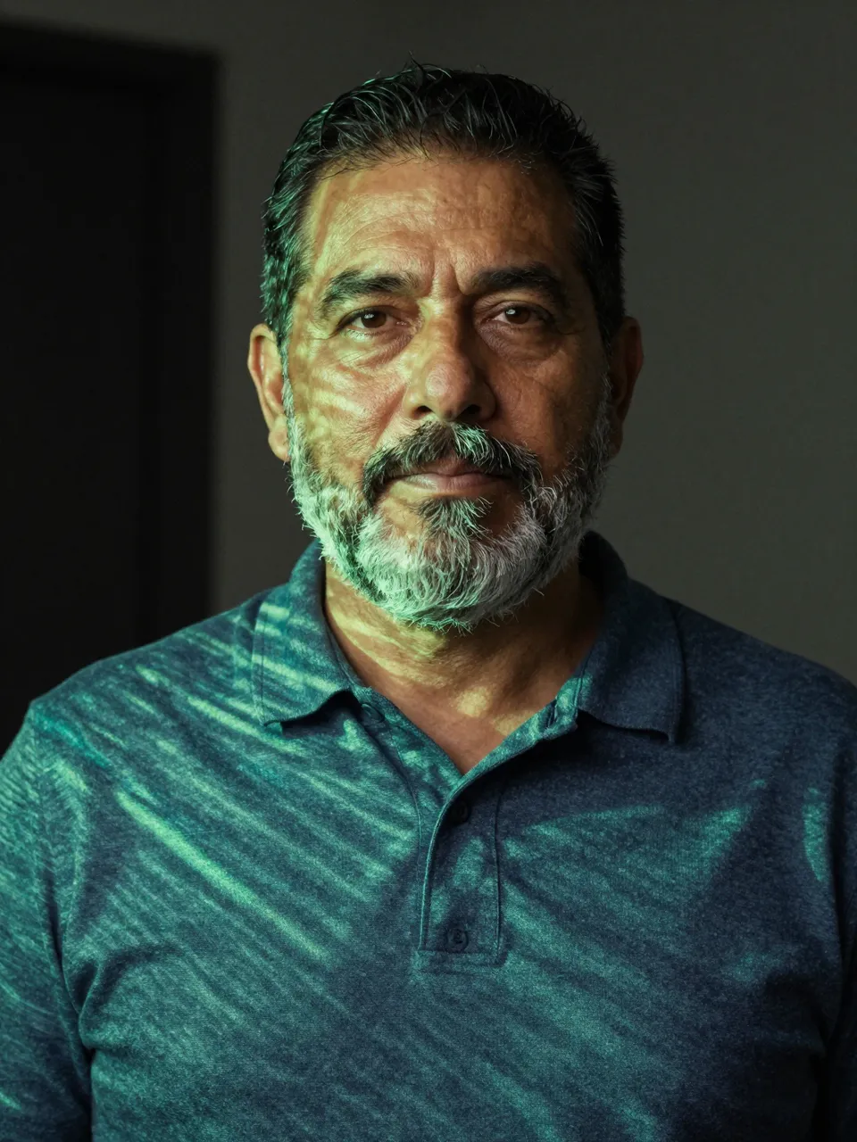 A sixties Latino man with a gray beard stands in a dark room lit by thin green projected lines with a calm expression
