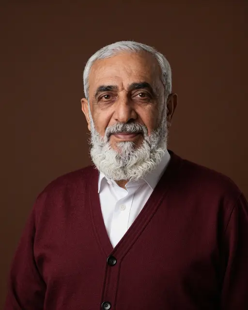 Studio portrait of a senior Middle Eastern man with a full white beard in a burgundy cardigan against a dark brown backdrop