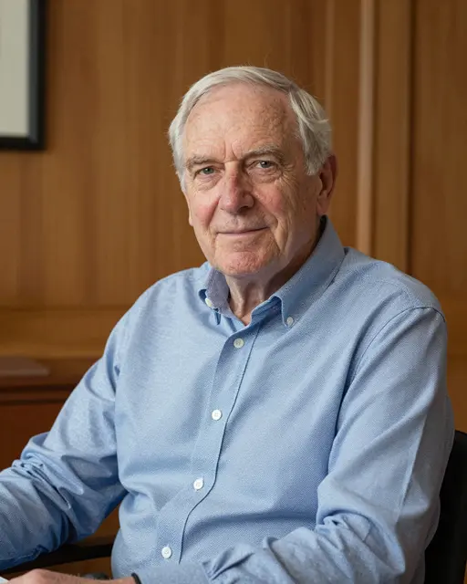 An elderly white man in his eighties sits looking at the camera in a wood-paneled study, wearing a light blue button-down