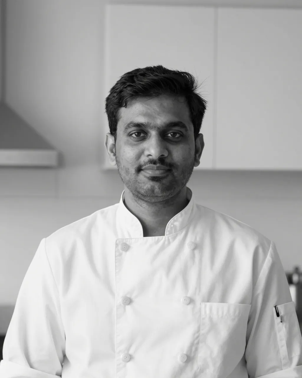 Black and white portrait of a South Asian man in a chef jacket looking at the camera in a kitchen