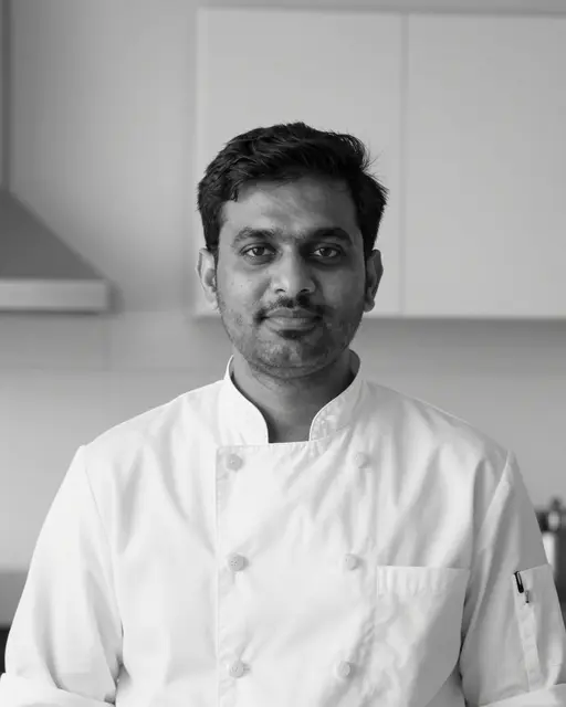 Black and white portrait of a South Asian man in a chef jacket looking at the camera in a kitchen