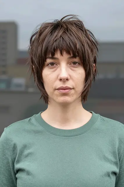 Woman in her thirties with a choppy shag haircut and green sweater stands facing the camera in overcast outdoor light
