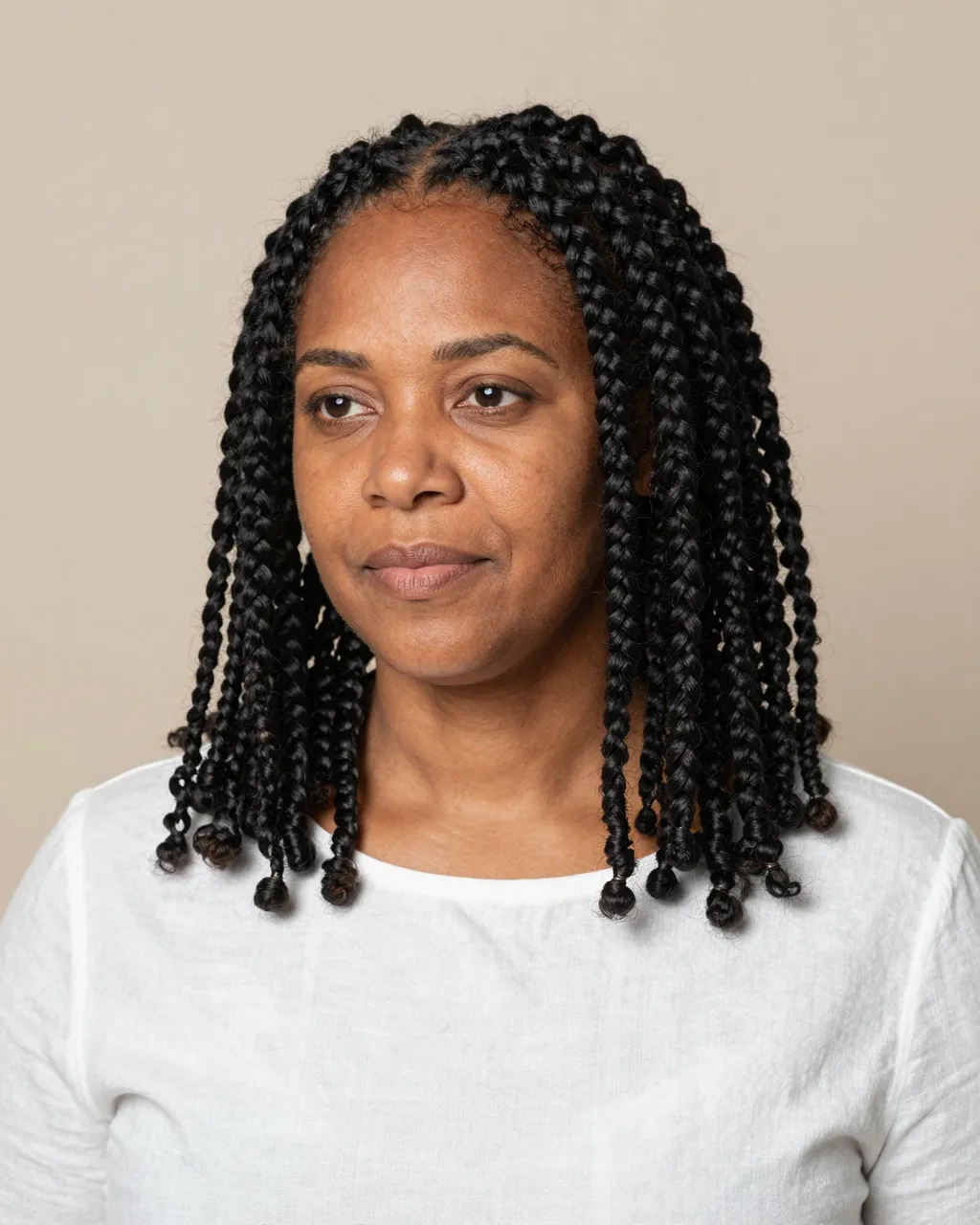 Studio portrait of a woman with shoulder-length braids wearing a white linen top against a beige background