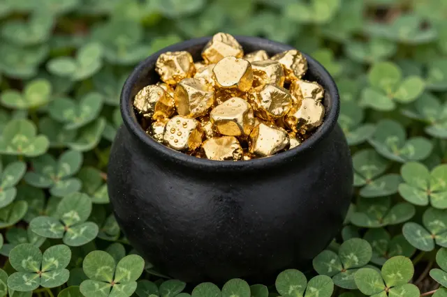 A small black pot filled with shiny gold nuggets sits on a bed of green clover leaves.