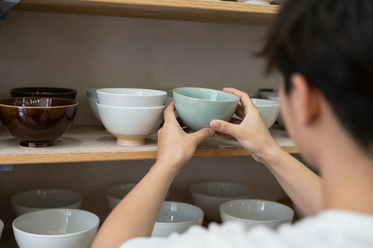 Seen from behind, a potter places a light blue glazed bowl on a wooden shop shelf of ceramics