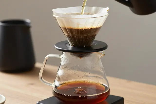 Hot water pours through a paper filter into a glass dripper above a clear coffee carafe on a wooden countertop.