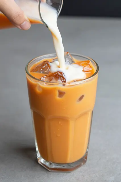 A hand pours cream from a small pitcher into a glass of orange Thai iced tea with ice cubes