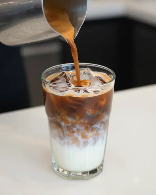 Espresso is poured from a metal pitcher into a clear glass of iced milk on a white countertop.