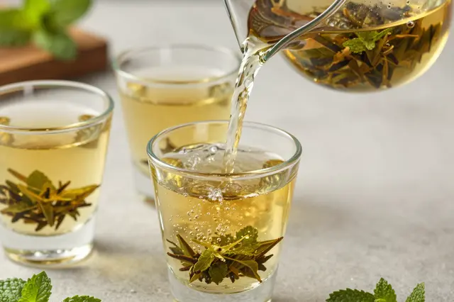 Golden mint tea is poured from a glass pitcher into small clear glasses on a light countertop