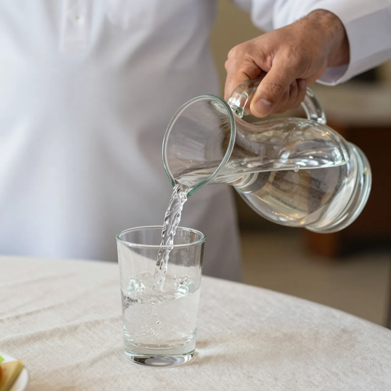 A hand pours clear water from a glass carafe into a drinking glass on a linen tablecloth